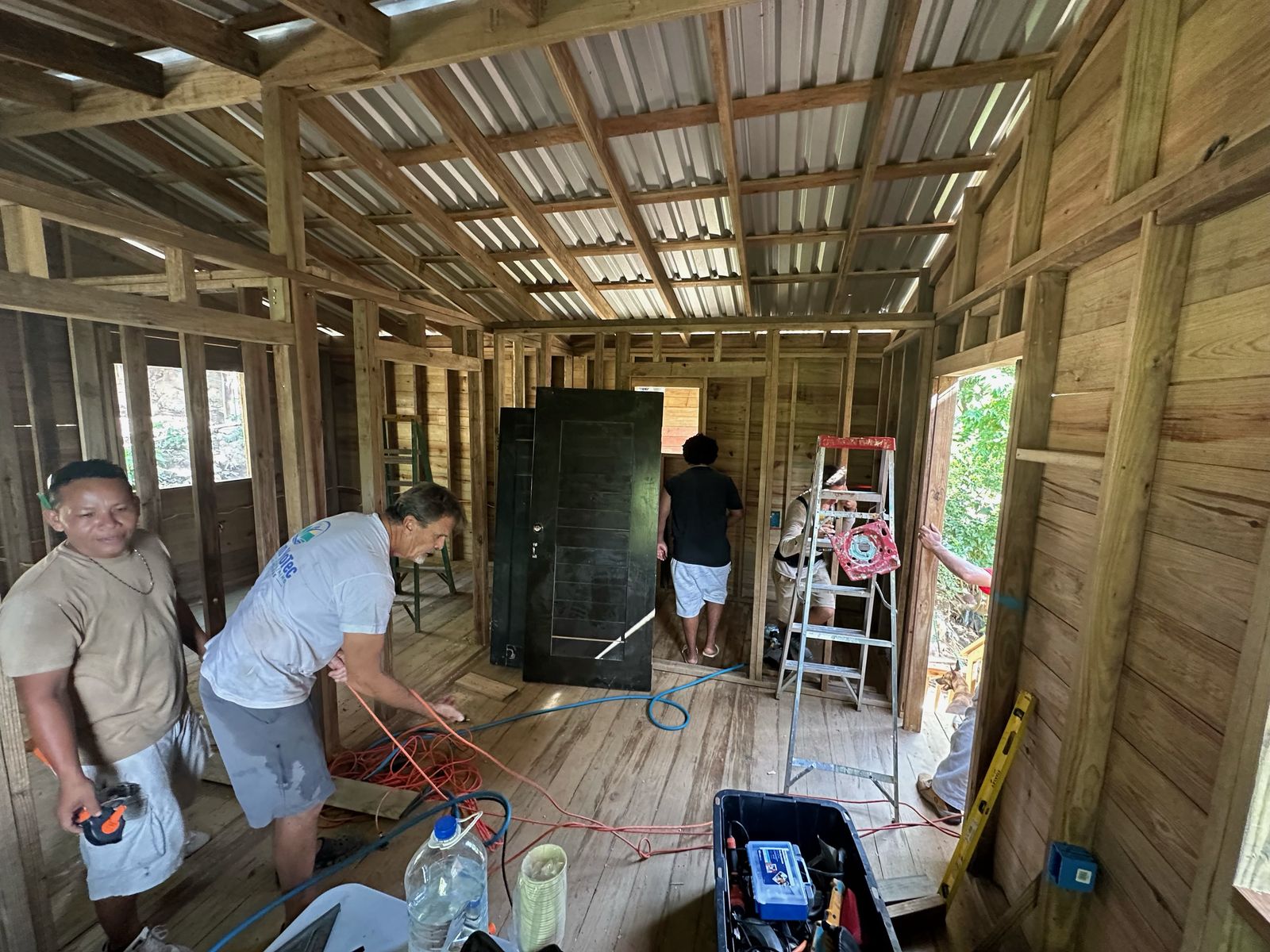 Two volunteers carry a dark wooden door through the framed home while others run wire
