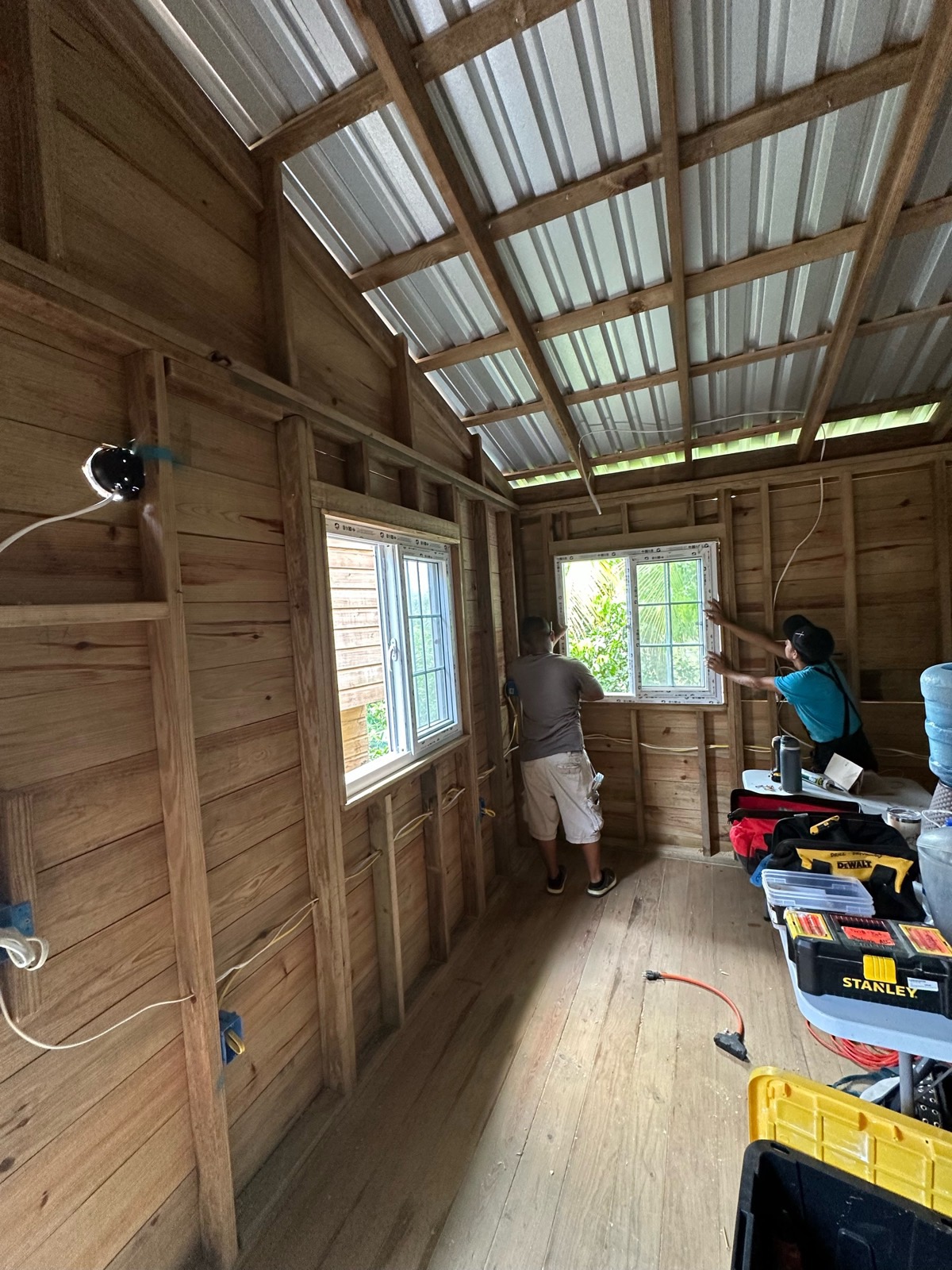 Two volunteers set a white window into the framed wall under the new metal roof, daylight through the trees outside