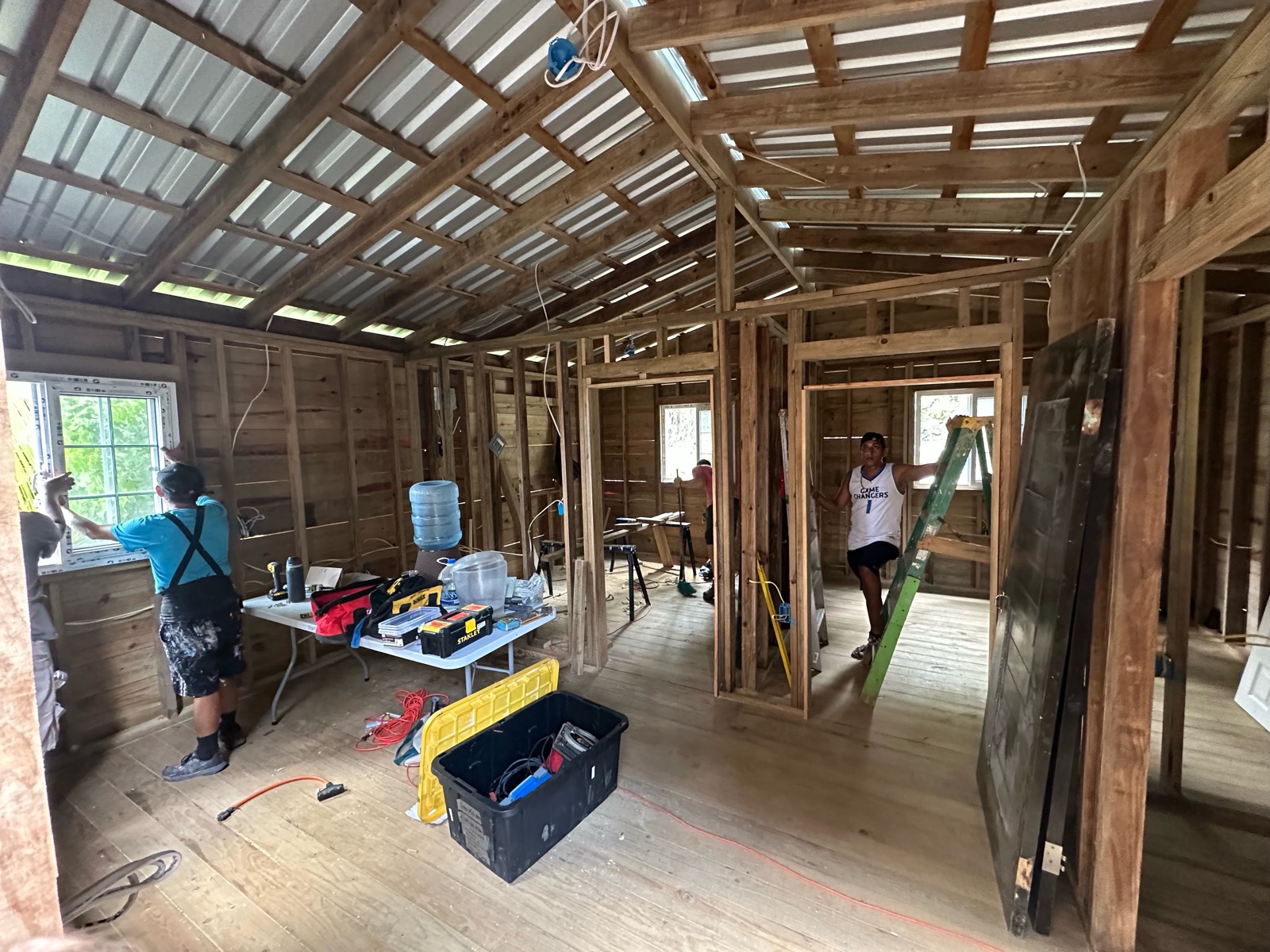 Inside the framed home under the new metal roof: a volunteer fits a white window on the left, a builder steadies a ladder in the bedroom doorway, dark doors lean ready to hang