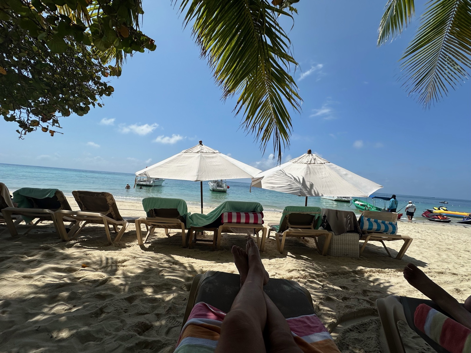 Lounge chairs and white umbrellas on a white sand beach under palm fronds, turquoise water and boats beyond